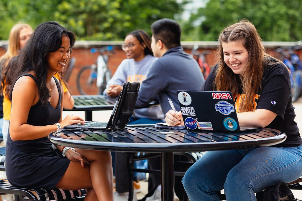 Two female students work on their laptops in front of the Zeanah Engineering Complex. 