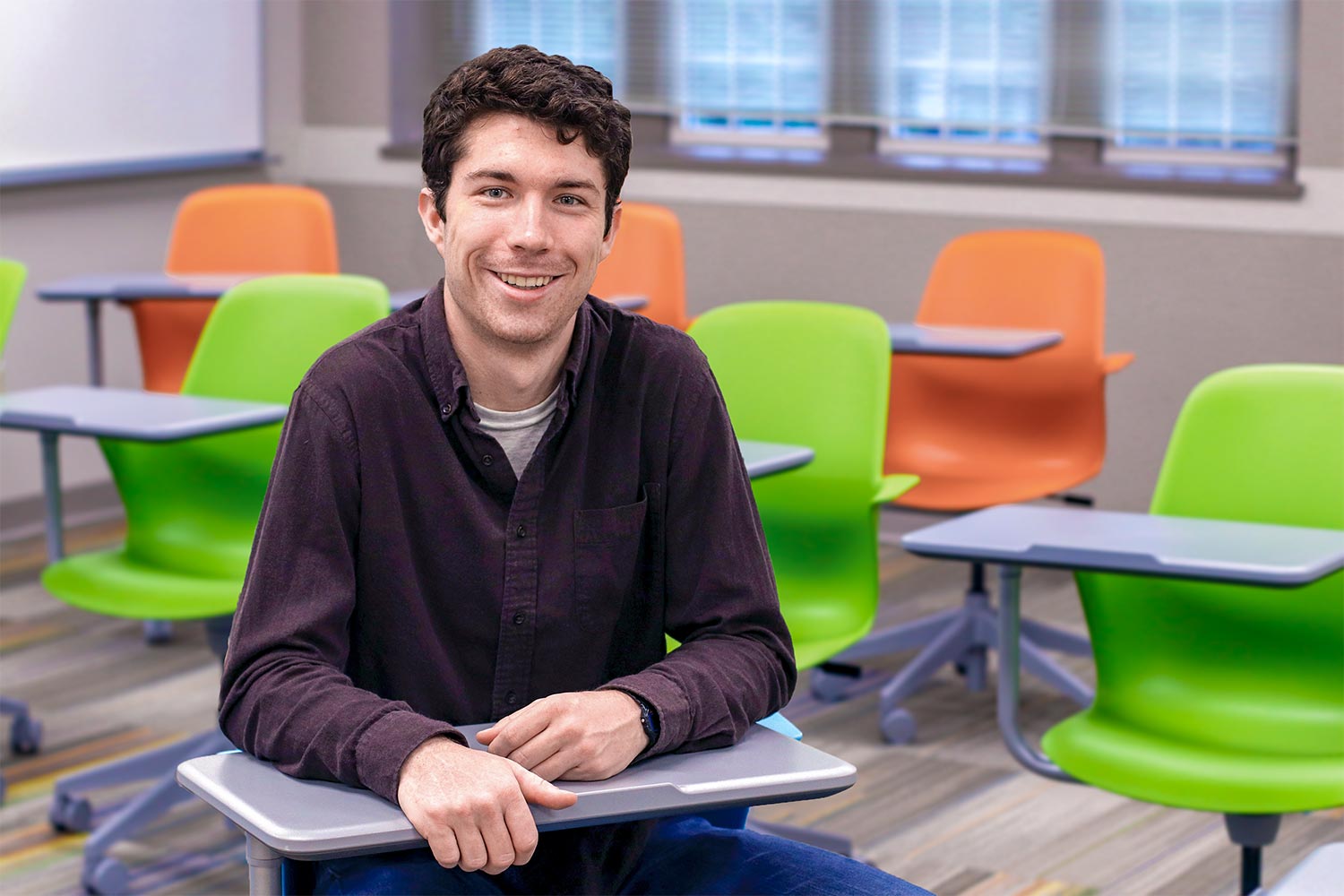 Eli Darby sits at a desk in a classroom, surrounded by other desks with bright orange and lime green seats.
