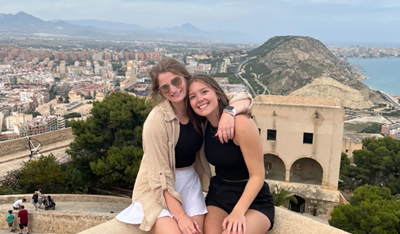Two women sit on a ledge above a city.