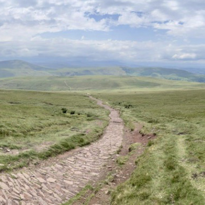 A view of the Pennine Way path that showcases the vastness of the trail highlighted by rolling hills and green grass with a cobblestone pathway. The Pennine Way path is mostly located in England with some parts stretching into Wales.