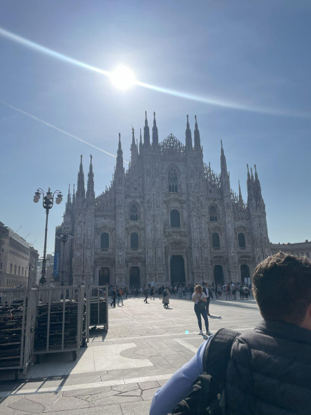 The historic site of Piazza del Duomo in Milan viewed from ground level. A tourist stands in the immediate foreground with their back to the camera and the large Piazza del Duomo building stands in the background with the sun brightly shining above it.