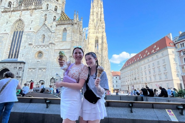 Cameron Castleberry and another study abroad student stand together in front of a cathedral style building while showing off ice cream cones. Both students are wearing cross body purses and white skirts.