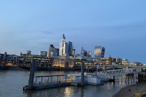 The a boat dock on the Thames river is shown in the foreground with a view of the downtown skyline of London, England in an early evening setting is shown in the background.