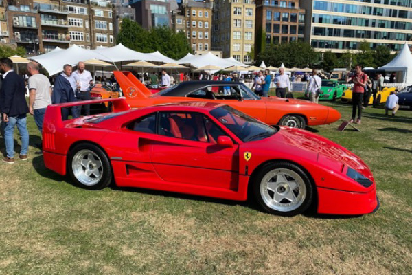 A red Ferrari F40 is shown in front of an orange 1970 Plymouth Cuda parked on grass at a car show in London, England.