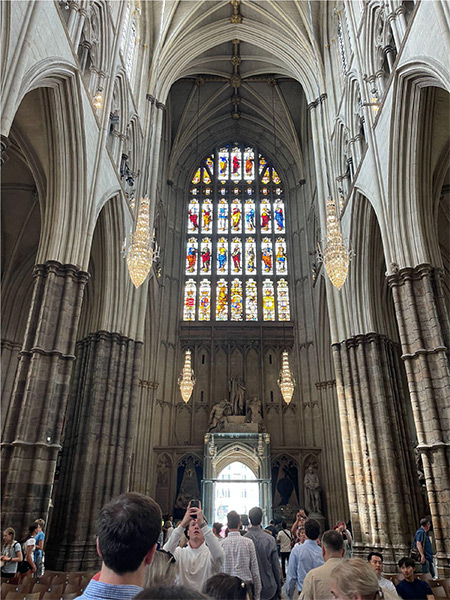 An interior view of Westminster Abbey. A tourist group walks among the inside of Westminster Abbey with a view of stained glass windows, archways, and decorative chandeliers hanging from the ceiling.