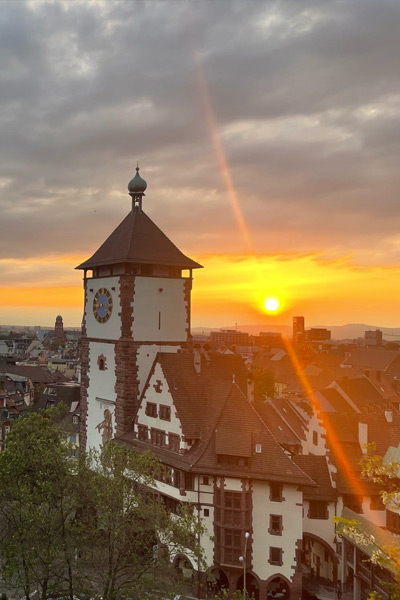 A vibrant sunset, accompanied by an orange sky with dark cloud cover, is viewed in the background of the Schwabentor building in Germany.