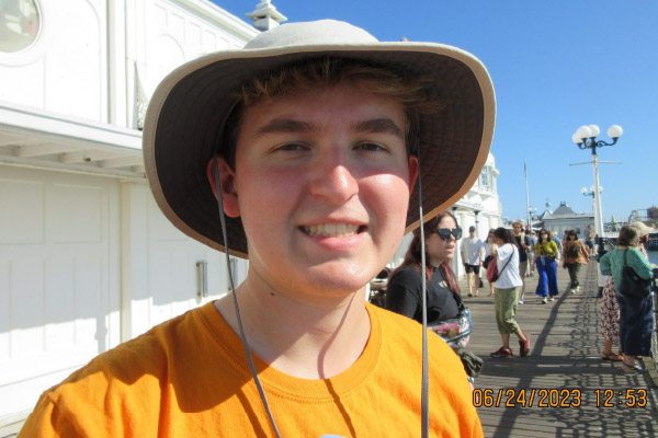 Jaxon Thornburgh smiles while standing on a boardwalk. Jaxon is dressed in a UT orange t-shirt and a tan wide brim hat.