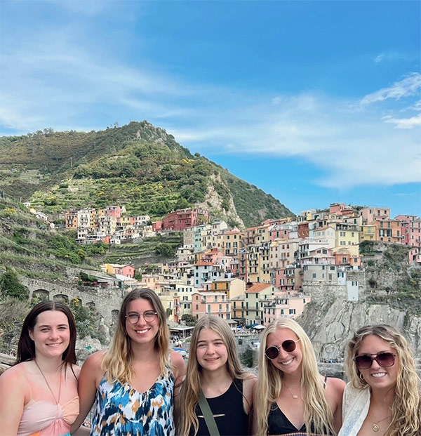 Madilyn Morelock and other study abroad engineering students pose together in a group at a location overlooking a town in Italy. The town in the background is densely populated with homes and buildings of varying shades of pink, yellow, green, and orange. 