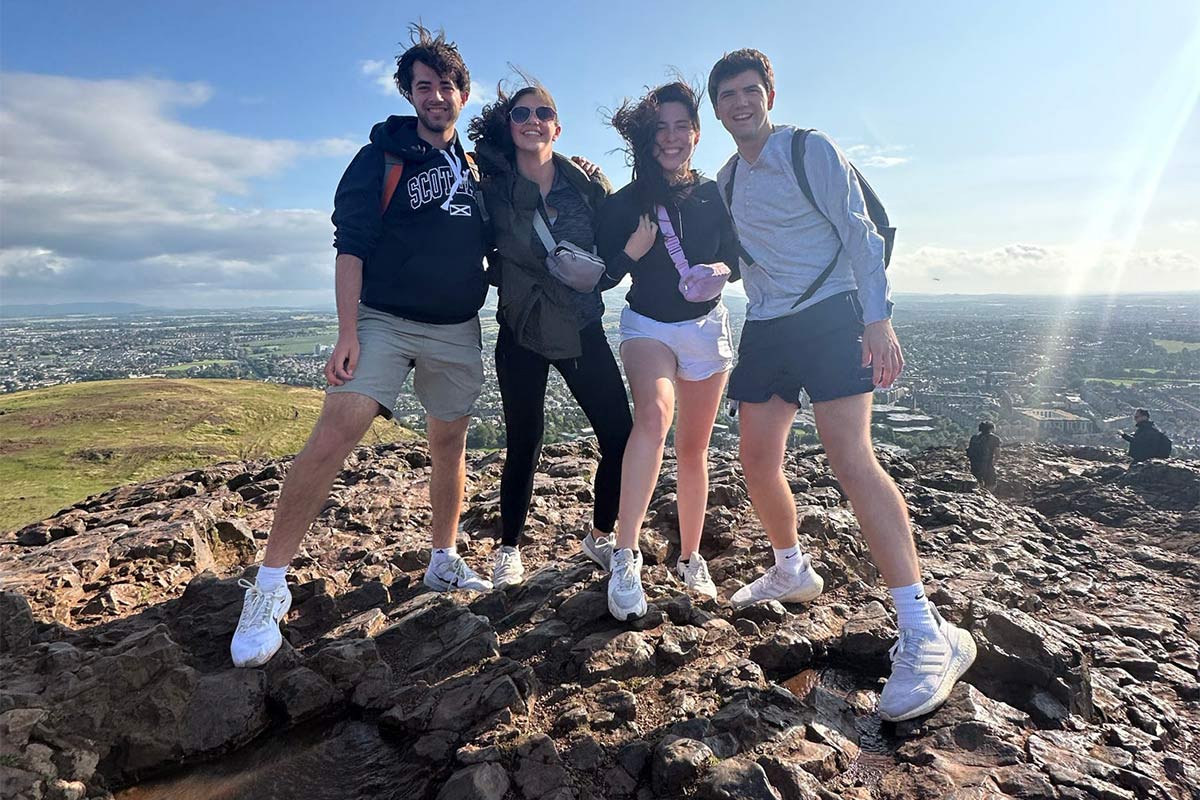 TCE student Mason Roddy and three other Engineering in London study abroad students stand together in a group with a blue sky background on top of a hillside that overlooks a large green field and cityscape.