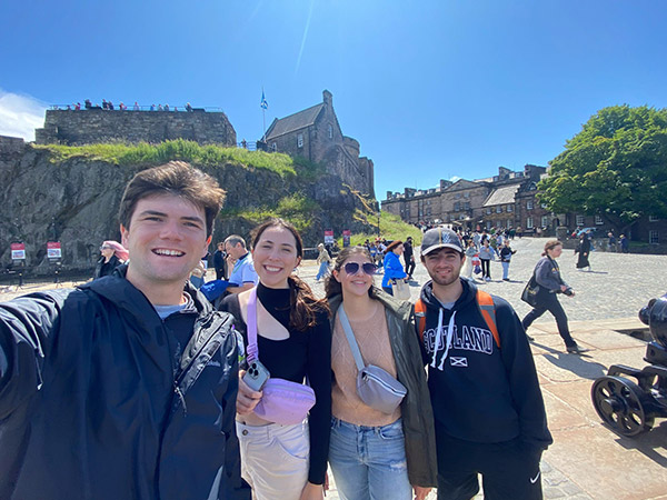 Mason Roddy and three other Engineering in London study abroad students standing in front of a cliff that is topped with green grass. This cliff is positioned in front of a castle-like building and a downtown shop area is shown to the right.