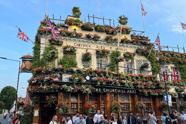 An exterior view of The Churchill Arms in London. A two-story building with multiple British flags along with plants and shrubs that adorn the top half of the buildings exterior.
