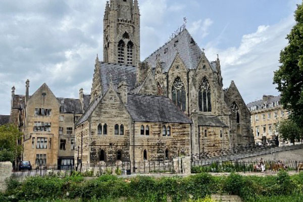 St. John's Roman Catholic church located in Bath, viewed from the ground level. The tan brick and cobblestone exterior appears aged and weather. The peak of the cathedral is not visible due to its height in relation to the rest of the building.