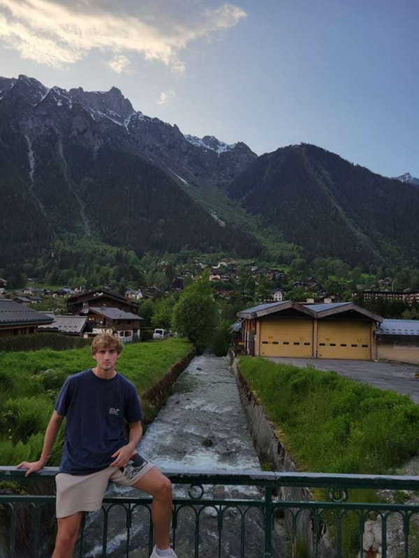Ryan Harper leans against a handrail of a bridge that overlooks a canal. Snow-capped grass covered mountains and a small village of homes are in the background.