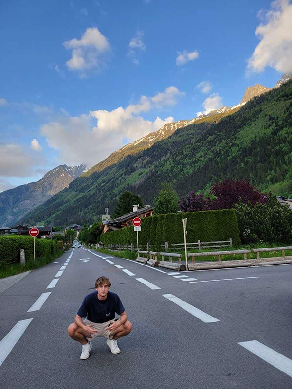 Ryan Harper squatting for a photo opportunity in the middle of a road. A blue sky highlights the large mountain range covered by trees and green plant life capped with snow that stands behind him.