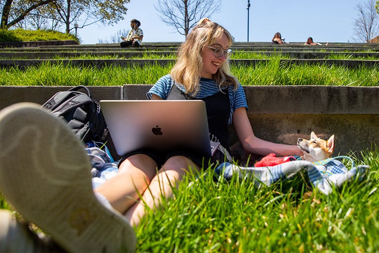 Student sits in grass with dog and laptop