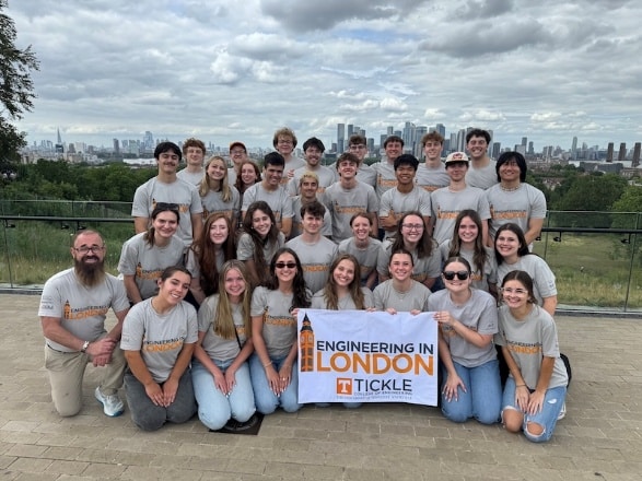 Study abroad cohort wearing matching gray t-shirts holding an "Engineering in London" banner with the city skyline behind them