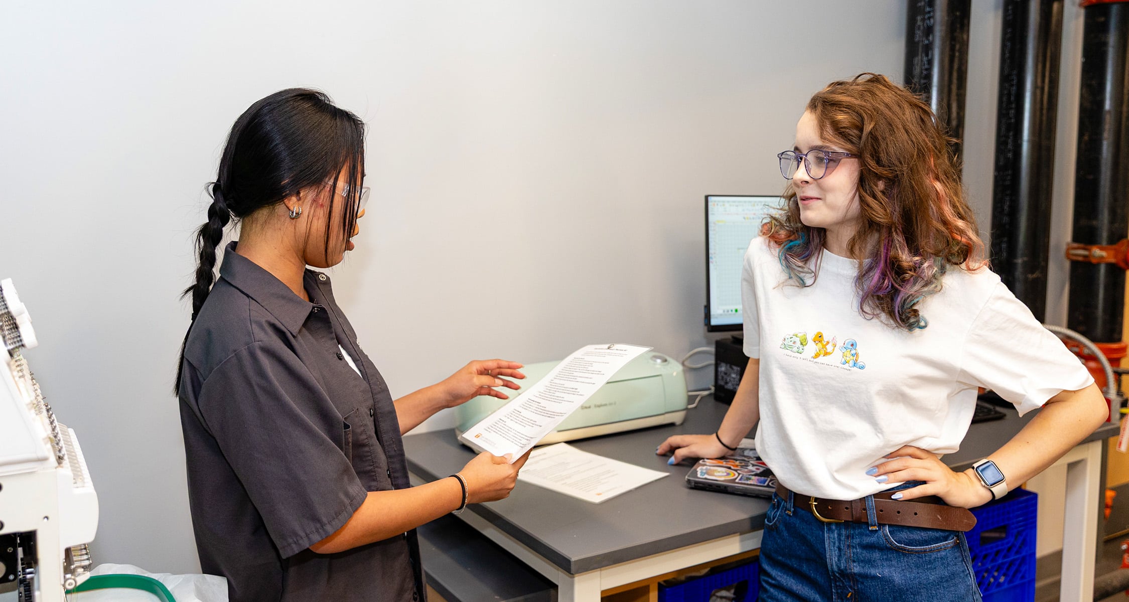 Two students stand and have a conversation in front of the digital sewing machine.